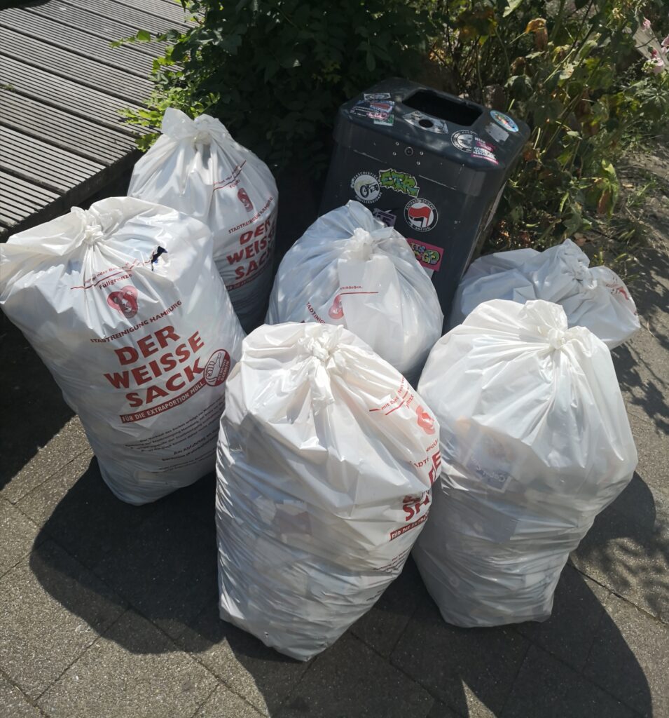 Sechs große weiße Müllsäcke stehen zur Abholung an einem Mülleimer der Stadt.